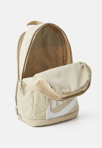 Beige stoffen rugzak met een afgeronde bovenkant, een voorvak met rits en een groot wit logodetail. De binnenkant is gevoerd met een lichter beige materiaal.
