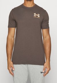 Brun kortärmad t-shirt i bomullsmix med en orange och grå Under Armour-logotyp på vänster bröst.