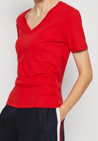 Kvinna som bär en åtsittande röd V-ringad T-shirt med korta ärmar och en hand i mörkblå byxor som har vit och röd sidosträng.