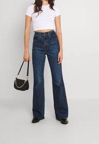 Vysoké tmavě modré zvonové džíny s hladkým denimovým povrchem. V kombinaci s přiléhavým bílým crop topem a malou černou kabelkou.