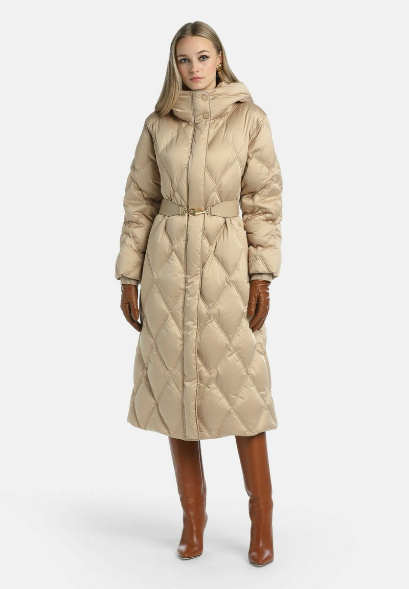 Puffjakke i beige med diamantquiltet mønster, høj krave, bælte i taljen og lange ærmer. Modellen bærer brune handsker og knæhøje støvler.