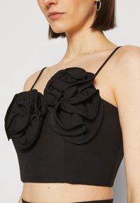 Femme portant un top court noir avec fines bretelles et grands détails floraux en tissu sur la poitrine et des boucles d'oreilles créoles argentées.
