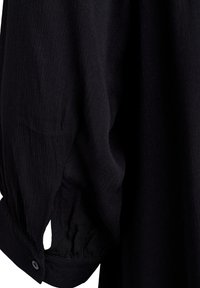 Manche longue noire avec poignet boutonné, tissu froncé créant un léger effet de manche bouffante.