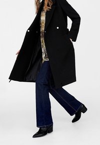 Abrigo de lana negro con diseño de doble botonadura y botones en tono plateado, llevado sobre una blusa de estampado y jeans de campana azul oscuro, con botas negras hasta el tobillo.