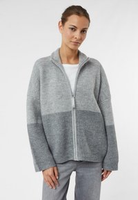 Grauer gerippter Reißverschluss-Cardigan mit einem color-block Design in helleren und dunkleren Farbtönen. Lange Ärmel und ein hoher Kragen. Weicher Stoff.