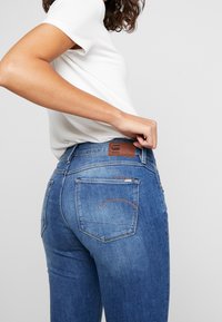 Jeans de denim azul con un diseño de tiro medio, costuras detalladas y un parche de cuero marrón en la cintura. Silueta ajustada y bolsillos traseros clásicos.