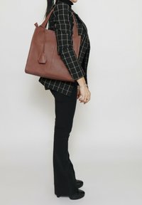 Bruine leren tote bag met een gladde textuur, rechthoekige vorm en zijaccentdetails. Gemaakt om over de schouder te dragen, gecombineerd met een zwart geruite blazer.