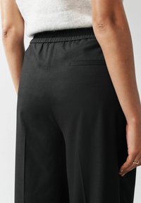 Person in schwarzen elastischen Taillenhosen mit einer Gesäßtasche, kombiniert mit einem weißen, flauschigen Oberteil, von hinten gesehen.