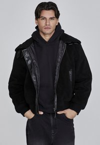 Chamarra de forro polar negra con acentos de cuero, cierre frontal y dos bolsillos laterales. Llevada sobre un sudadera negra con capucha, cuenta con un cuello tipo solapa y tejido texturizado.