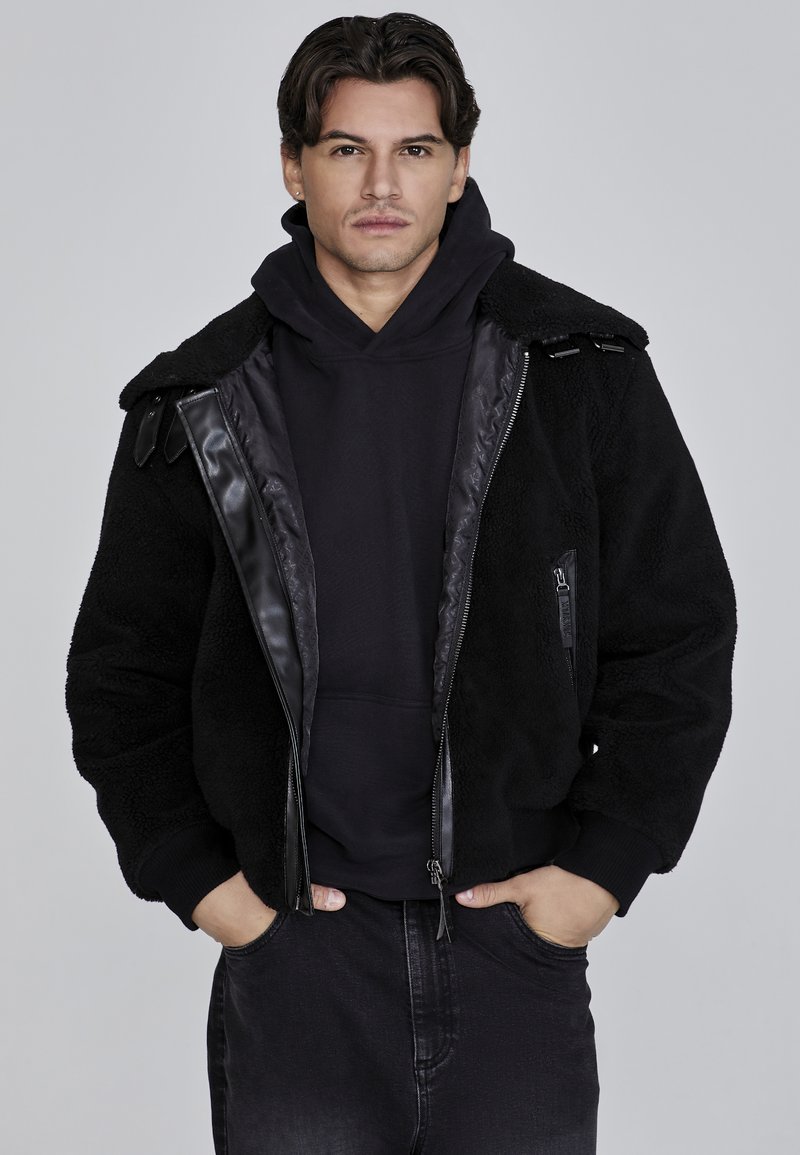 Chamarra de forro polar negra con acentos de cuero, cierre frontal y dos bolsillos laterales. Llevada sobre un sudadera negra con capucha, cuenta con un cuello tipo solapa y tejido texturizado.