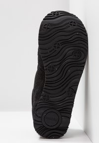 Černá teniska s texturovaným svrškem a gumovou podrážkou se zvlněným vzorem a kruhovými akcenty. Logo značky je viditelné na podrážce.