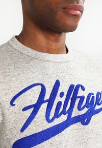 Szary sweter z heatherowym wykończeniem, z okrągłym dekoltem, z dużymi, teksturowanymi niebieskimi literami "Hilfiger" wyraźnie umieszczonymi na klatce piersiowej.