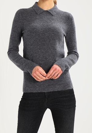 Femme portant un pull ajusté gris foncé à manches longues avec un petit col et un pantalon noir, les mains légèrement jointes devant.