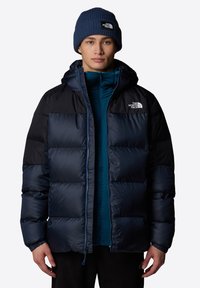 Niebieska kurtka puchowa z kapturem, z pikowanym wypełnieniem, czarnymi akcentami i zapięciem na zamek. Noszona na niebieskiej bluzce w wzory, z pasującą czapką.