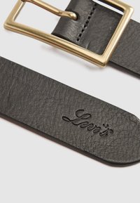 Gros plan sur une ceinture en cuir noir texturé avec une boucle en or rectangulaire et le logo Levi's en relief à l'extrémité de la sangle.