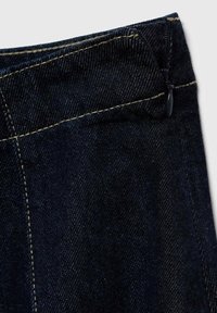 Tissu en denim foncé avec une texture lisse et des coutures contrastantes de couleur marron clair. Présente une fermeture éclair et un design de poches en biais à la taille.