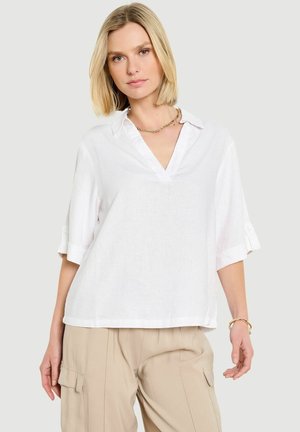 Blondynka w luźnej białej bluzce z podwiniętymi rękawami, beżowych spodniach cargo, z złotym naszyjnikiem i bransoletką, stojąca na jednolitym tle.