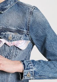 Džínová bunda s modrým práním, sklápěcím límcem, přední kapsou s kovovým knoflíkem a vzorovaným šátkem, který vykukuje zespodu.