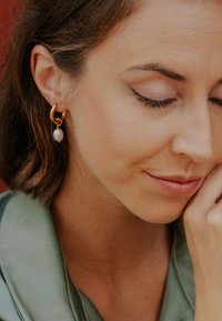 Cercei de tip cerc din aur cu un mic pandantiv de perlă. Tonul pielii moi al modelului și bluza verde din satin completează nuanțele calde ale bijuteriilor.