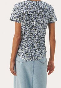 Mujer vistiendo una camisa floral de manga corta con flores azules, negras y blancas, combinada con una falda de mezclilla azul claro, vista desde atrás.