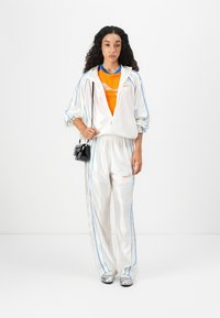 Tuta di satin bianco con strisce blu e logo arancione, indossato sopra una maglietta arancione. Il modello tiene una piccola borsa nera e indossa scarpe argentate.