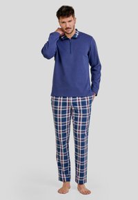 Camisa de manga larga azul marino con cuello de cremallera, combinada con pantalones de cuadros azules que presentan acentos en blanco y rojo. Tejido suave, corte relajado.