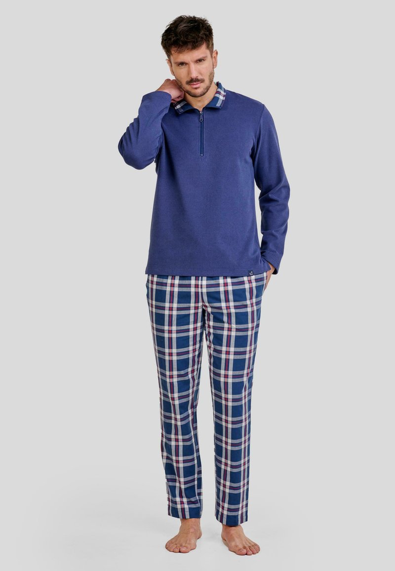 Camisa de manga larga azul marino con cuello de cremallera, combinada con pantalones de cuadros azules que presentan acentos en blanco y rojo. Tejido suave, corte relajado.