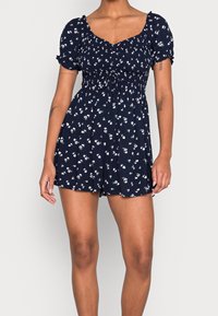 Navy blue floral print romper with short puff sleeves, a sweetheart neckline, and a cinched waist, featuring a relaxed fit and short shorts.