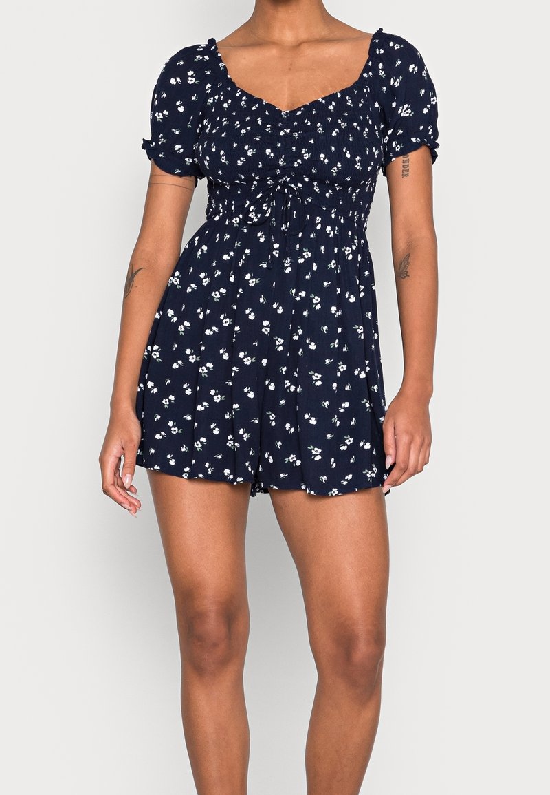 Navy blue floral print romper with short puff sleeves, a sweetheart neckline, and a cinched waist, featuring a relaxed fit and short shorts.