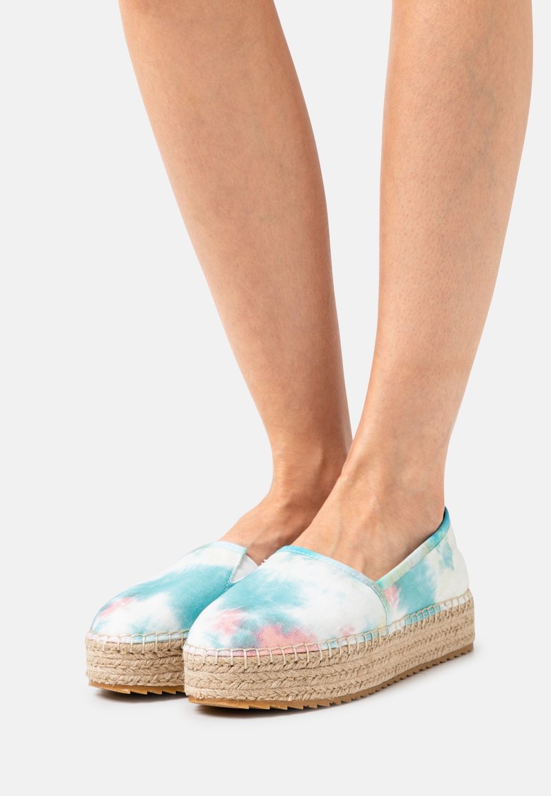 Jambes portant des espadrilles compensées en tissu tie-dye bleu, rose et blanc avec des semelles en jute tissée sur un fond clair.