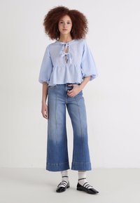 Blusa a scacchi blu chiaro con un nodo frontale, maniche a sbuffo e vita arricciata; abbinata a jeans in denim blu a gamba larga e scarpe nere.