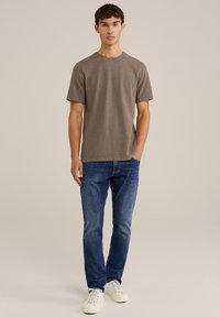 Tricou din bumbac maro cu mâneci scurte, gât rotund, purtat cu blugi albaștri și adidași albi. Design simplu și lejer.
