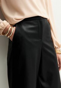 Pantalones de cuero negro con un diseño limpio, que cuentan con un bolsillo lateral y una textura suave. Combinados con una blusa de durazno claro y pulseras de oro.