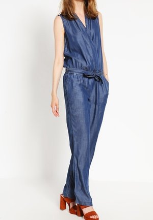 Jeans-Jumpsuit mit Wickelvorderseite, ärmellosem Design, elastischem Taillenbund und seitlichen Taschen. Kombiniert mit braunen Sandalen mit Blockabsatz.