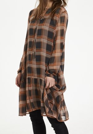 Kvinde iført en løs plaidkjole med lange ærmer i sort, orange og beige, holder stoffet med den ene hånd mod en ensfarvet lys baggrund.