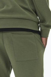 Sportowe spodnie w oliwkowym zielonym kolorze o miękkiej fakturze, z tylną kieszenią i czarnym pasem w talii. Prosty design podkreśla wygodę i funkcjonalność.