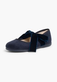 Zapato de ante azul marino con suela plana y un amplio lazo de terciopelo en la parte superior. El interior es de un color claro y texturizado.