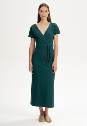 Vrouw staat en draagt een donkergroene korte mouw wikkeljurk met een strikceintuur en beige gevlochten platte schoenen, neutrale achtergrond.