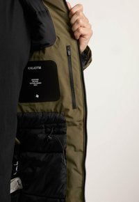 Puffermantel mit olivgrüner Außenseite, schwarzem Innenfutter, Reißverschluss-Tasche und Markenetikett. Verstellbarer Kordelzug in der Tasche.