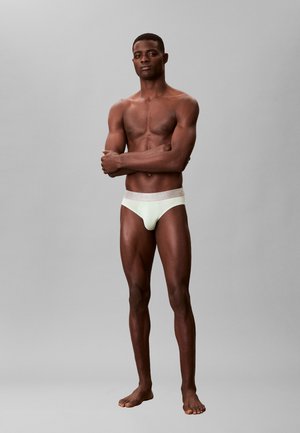 Uomo muscoloso in piedi a piedi nudi con le braccia incrociate, indossando slip chiari su sfondo grigio neutro.