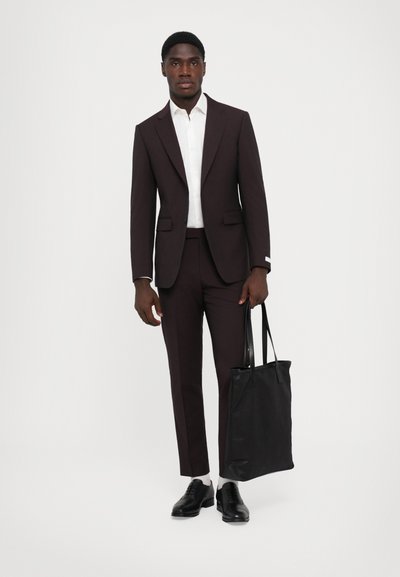 Completo tailored marrone scuro, camicia bianca e scarpe nere. Tiene una borsa nera e sta in piedi su uno sfondo bianco.