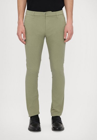 Pantalon vert clair en tissu lisse, à coupe slim, avec des passants de ceinture et une fermeture éclair à l'avant. Porté avec des chaussures noires.