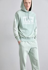 Mintgrøn sweatshirt med snøre, med en frontgrafik og tekst på ærmerne. Sammen med matchende sweatpants, glat tekstur.