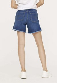 Shorts en jean de couleur bleu foncé avec un ourlet effiloché, dotés de deux poches arrière et de coutures visibles. Accompagnés de baskets blanches.