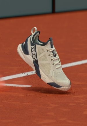 Scarpa da tennis K-Swiss beige e blu navy sospesa in aria sopra un campo di terra rossa, con particelle di terra rossa che si disperdono vicino alle linee bianche del campo.