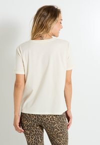 Mujer con cabello rubio que lleva una camiseta de manga corta color crema lisa y pantalones con estampado de leopardo, de espaldas a la cámara contra un fondo blanco.