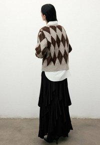 Mujer con cabello largo y oscuro que lleva un suéter de patrón de diamante marrón y beige, camisa blanca y una larga falda negra con volantes, frente a una pared blanca.