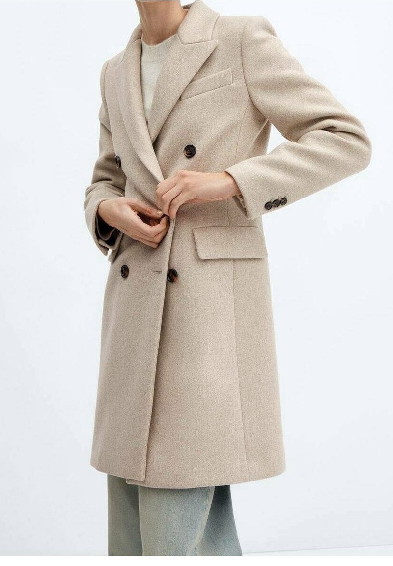 Beige dobbeltbrystet frakke lavet af uldblanding med spidse lapeler, to forlommer og brune knapper, parret med lyse denimbukser.