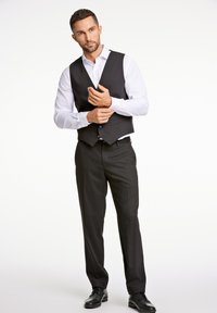 Gilet nero sopra una camicia bianca, abbinato a pantaloni e scarpe neri. Presenta una vestibilità su misura con bottoni e una texture liscia.
