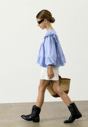 Blouse bleu clair à volants avec des bordures en dentelle, jupe fleurie blanche, bottines en cuir noir jusqu'à la cheville et sac en osier avec des accents marron.